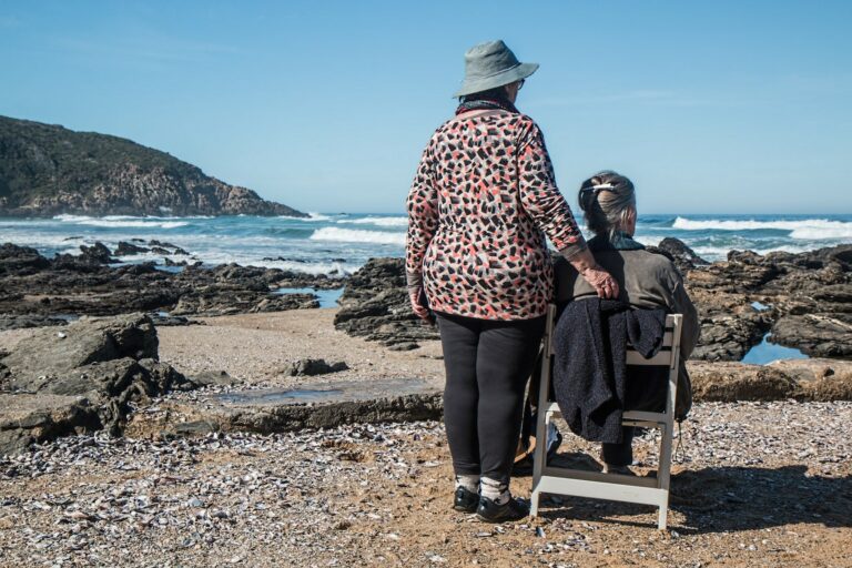 Two elderly people on the beach looking out towards the water; one is standing and one is seated